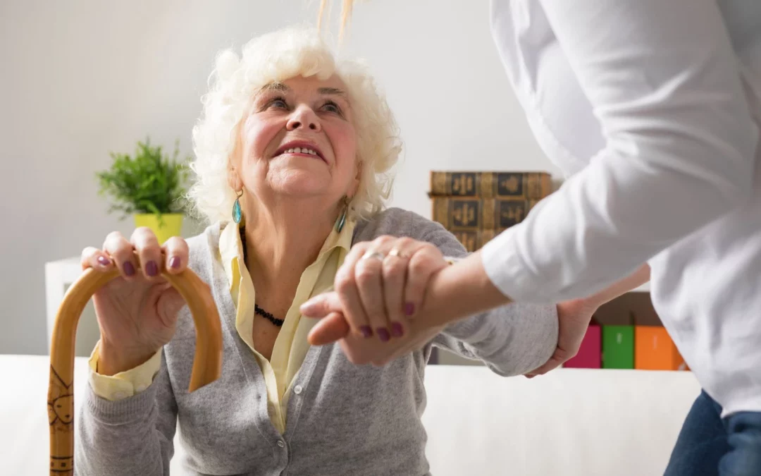 Auxiliaire de vie en CESU tient la main d’une dame âgée, assise sur un canapé, qui est son particulier employeur, afin de l’aider à se lever.