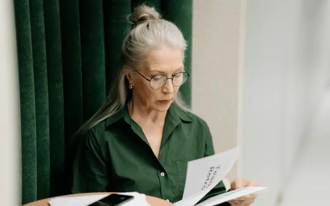 Femme senior aux cheveux gris, lunettes rondes, chemise verte, lisant attentivement un document administratif — illustre une aide à domicile en CESU qui prépare sa retraite.